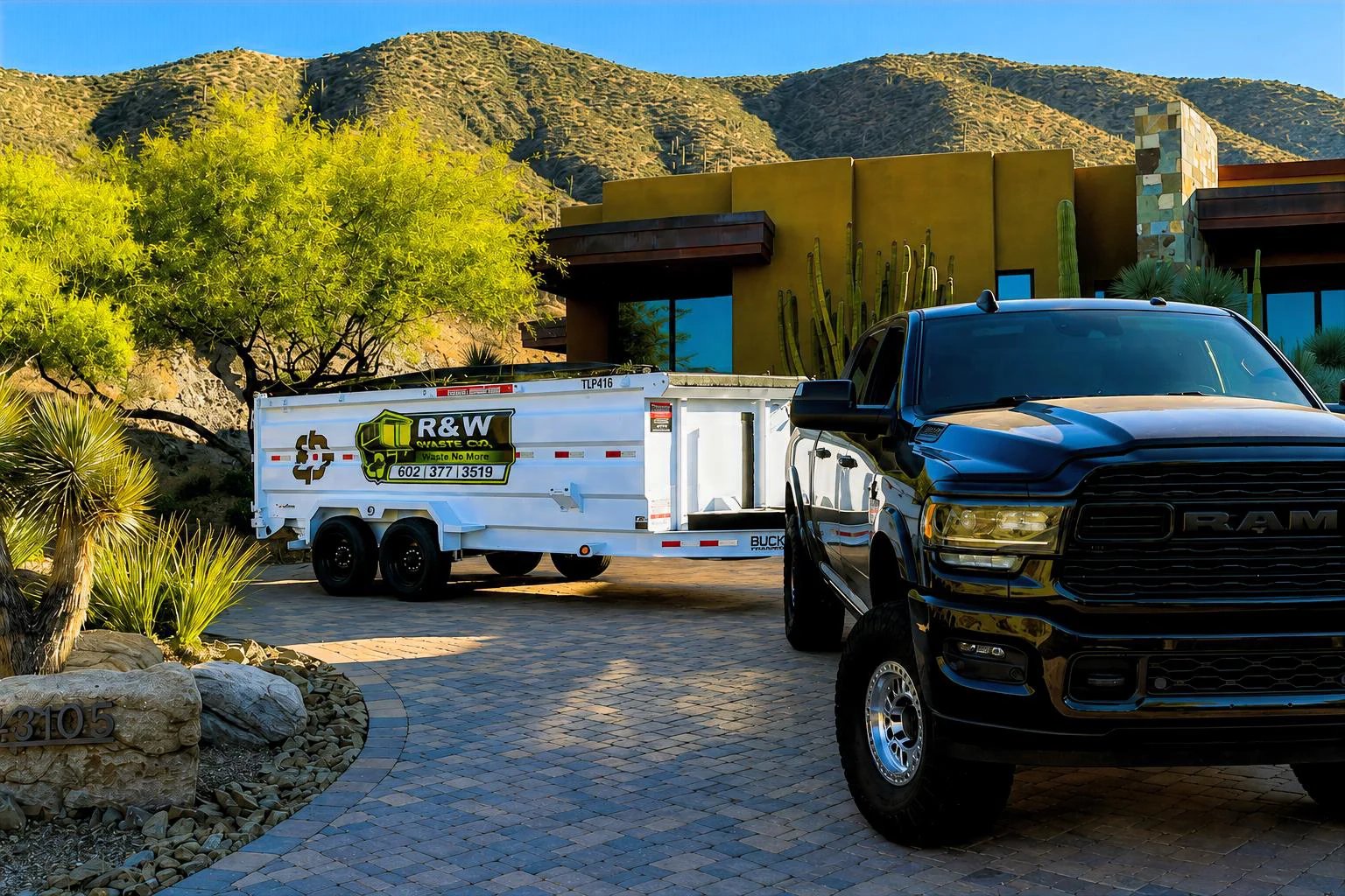 Black pickup truck parked next to white R&W utility trailer in desert driveway with modern home and yellow flowering trees